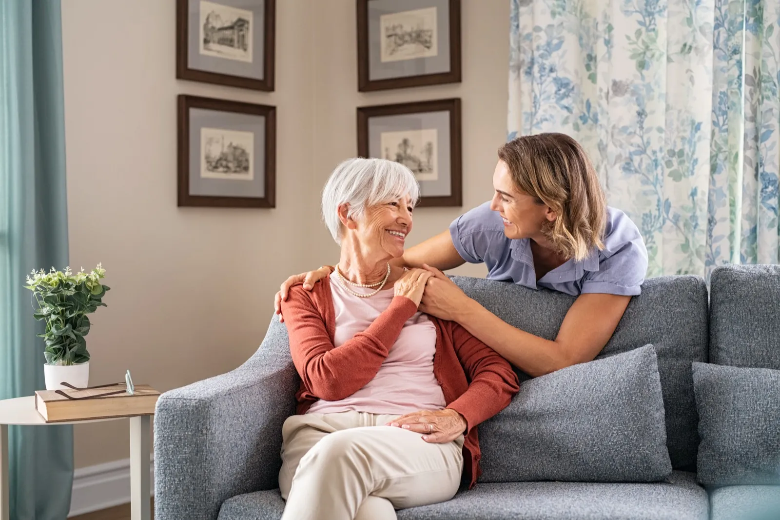 A caregiver leaning over the couch to embrace a smiling older woman at home