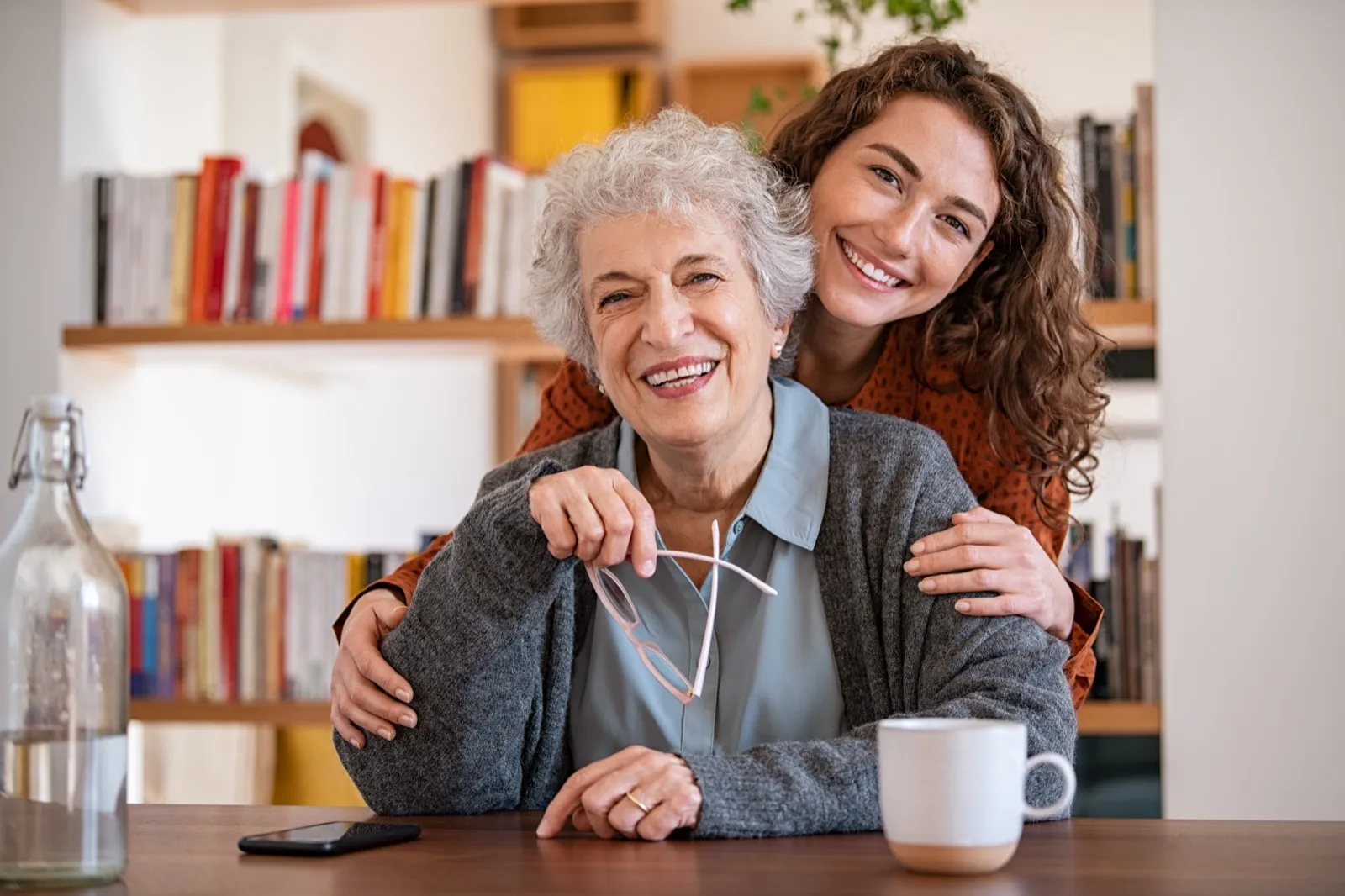 A grandmother at home smiling as her granddaughter hugs her from behind