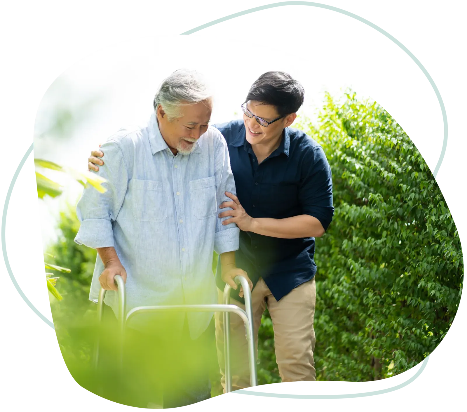 A younger man supporting an older man walking with a walker in a sunny garden