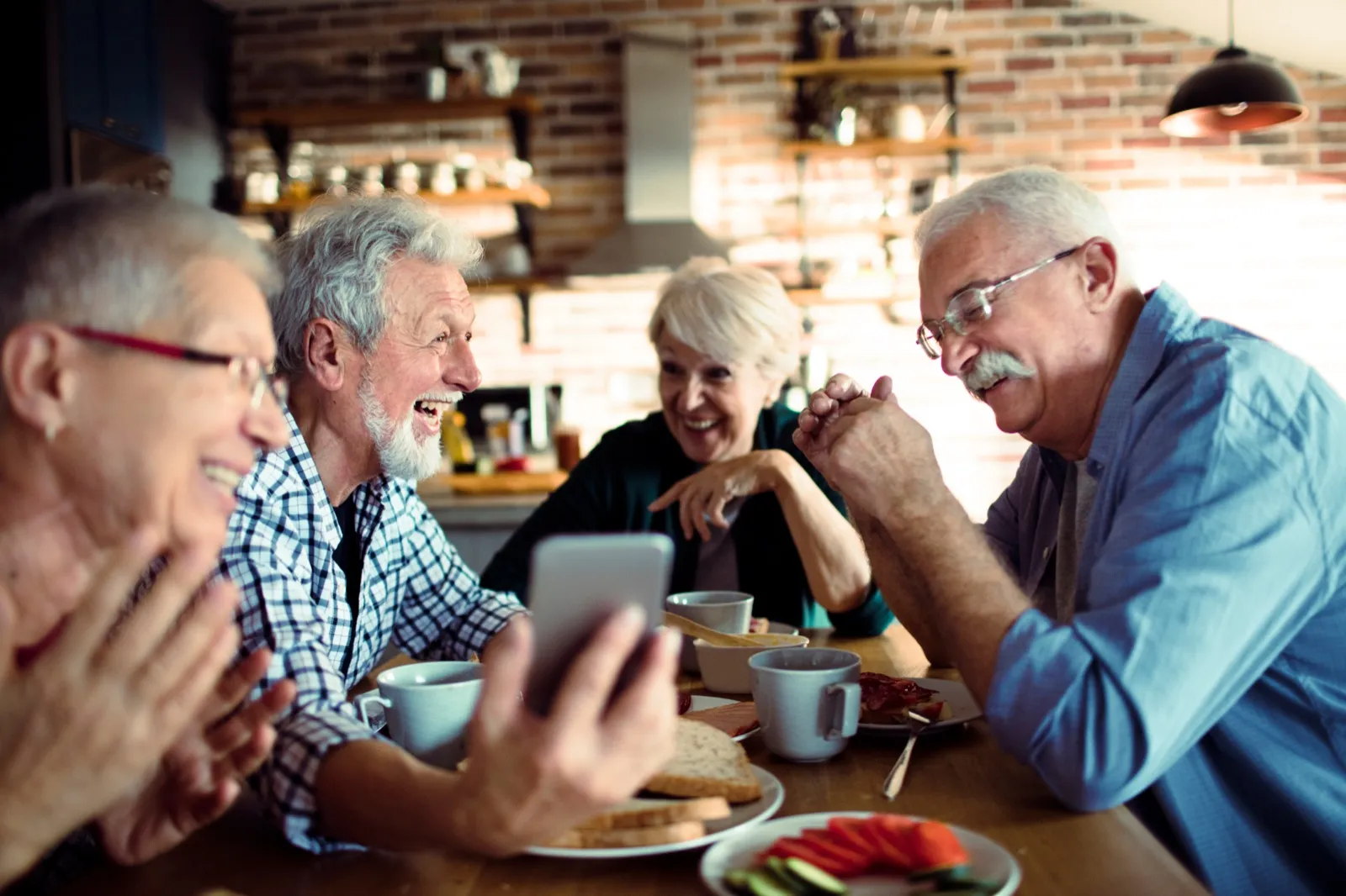Four older friends laughing together around a kitchen table