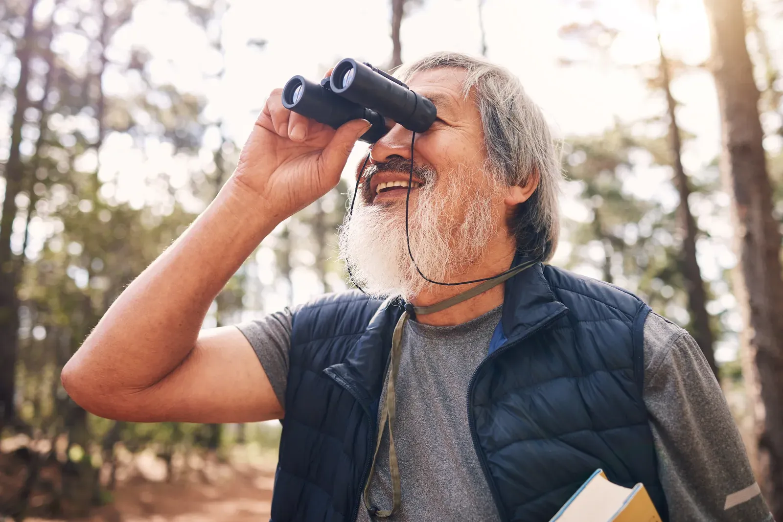 A smiling older man birdwatching through binoculars in a sunny forest