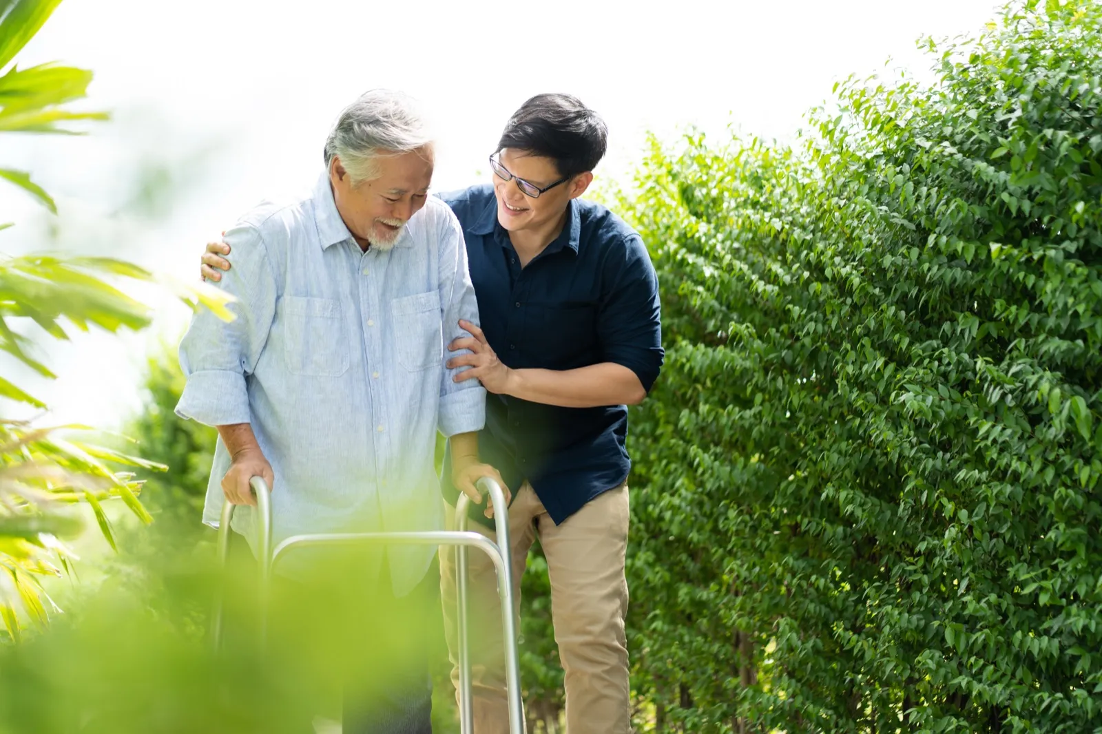 A younger man supporting an older man walking with a walker in a sunny garden