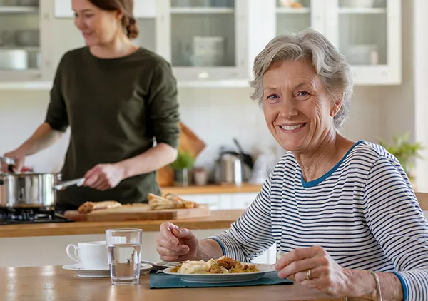 An older woman enjoying a meal while a Goldy Enrichment Specialist cooks in the kitchen