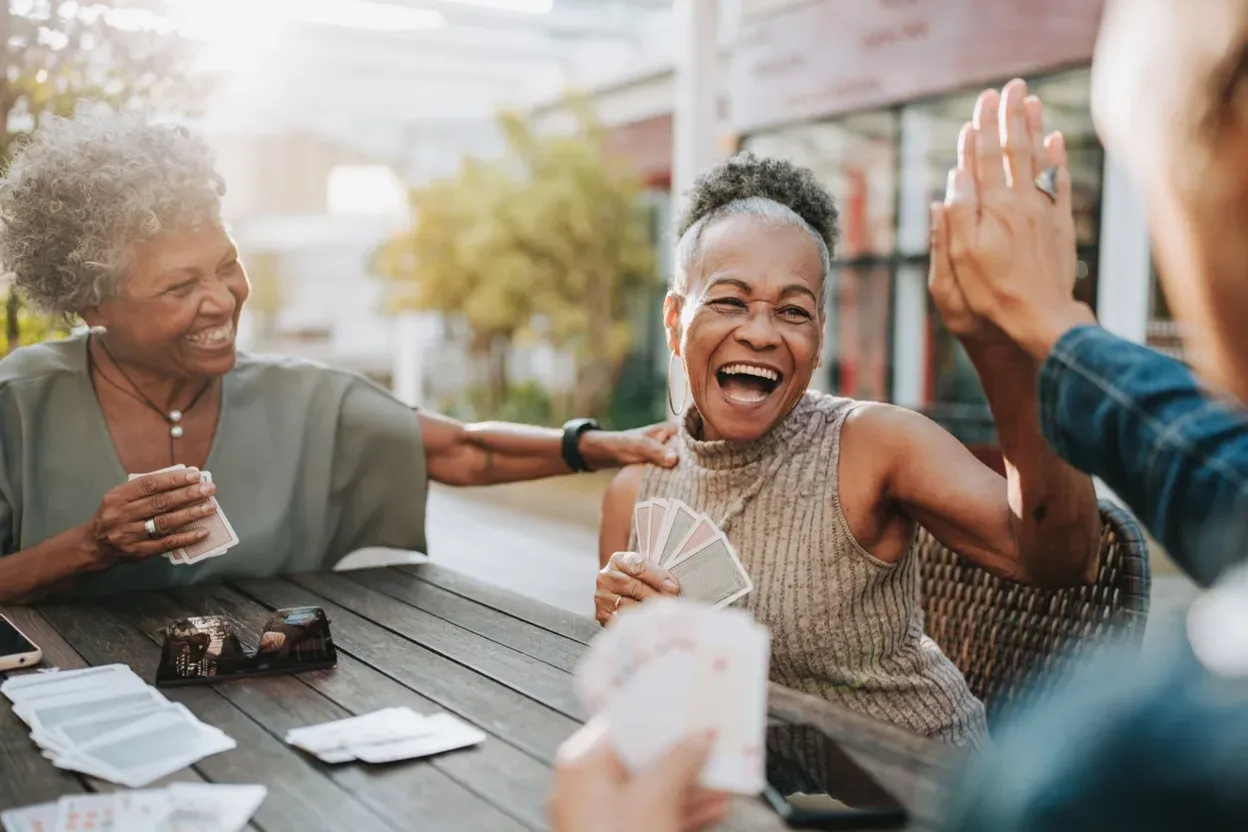A group of older friends laughing and high-fiving over a card game at an outdoor table